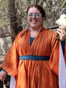 Laurencia wearing an orange flowing dress, holding a spindle and distaff