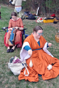 Two sitting women in medieval costume. One is weaving a band on a small loom, the other is spinning yarn on a drop spindle.
